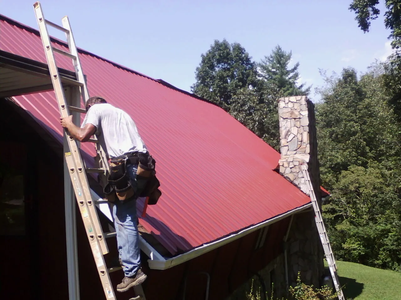 Roofer on a ladder beside a red metal roof and stone chimney — Soffit & Fascia Repair by Roofing Brothers in Moran